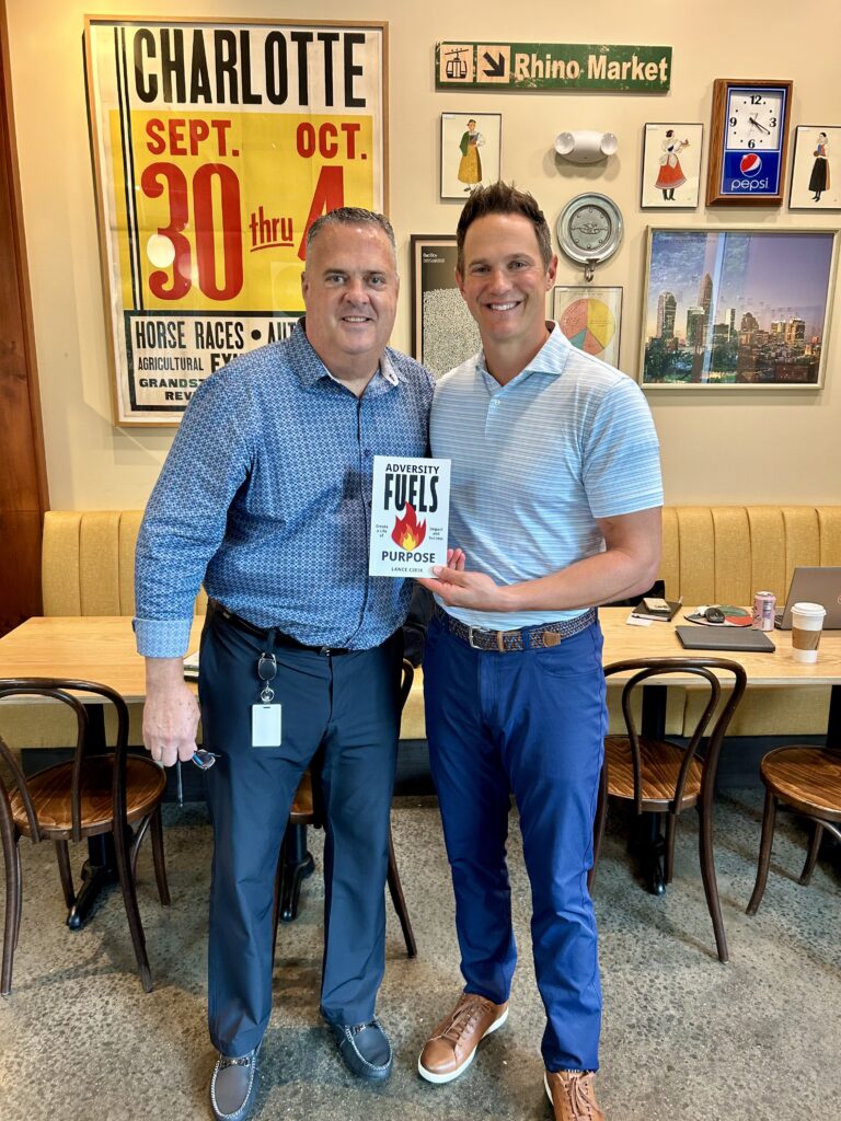 Lance Cibik standing with another person at Rhino Market in Charlotte, holding his book Adversity Fuels Purpose.