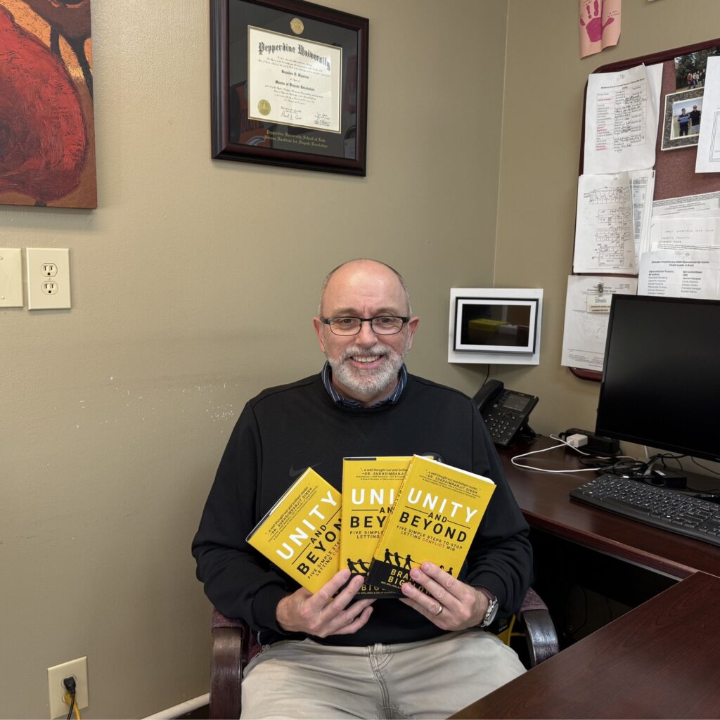 Photo of Brandon smiling in his office holding three copies of Unity and Beyond, his conflict resolution book, with his Pepperdine University dispute resolution degree visible on the wall behind him.