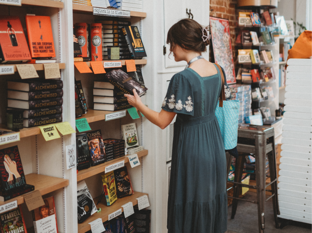Woman browsing books at an independent bookstore, pulling a title from a shelf lined with staff picks and handwritten recommendation tags.