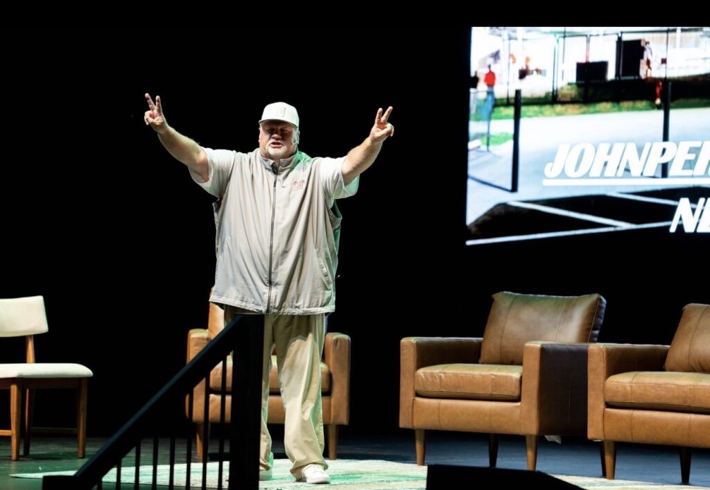 Photo of John Perry, football coach and author, speaking on stage with arms raised, personal growth book for coaches.