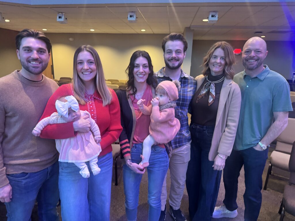 Photo of David Hockman smiling with his wife Sherry and adult children holding babies in a church gathering space, author of Surgeon Unmasked.