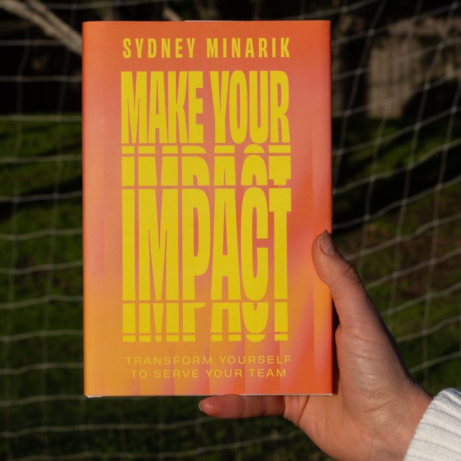 Sydney Minarik holding her debut book Make Your Impact Transform Yourself to Serve Your Team in front of a soccer goal net