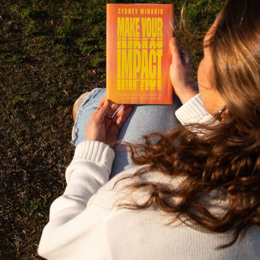 Sydney Minarik sitting on grass in golden hour light reading her debut book Make Your Impact Transform Yourself to Serve Your Team