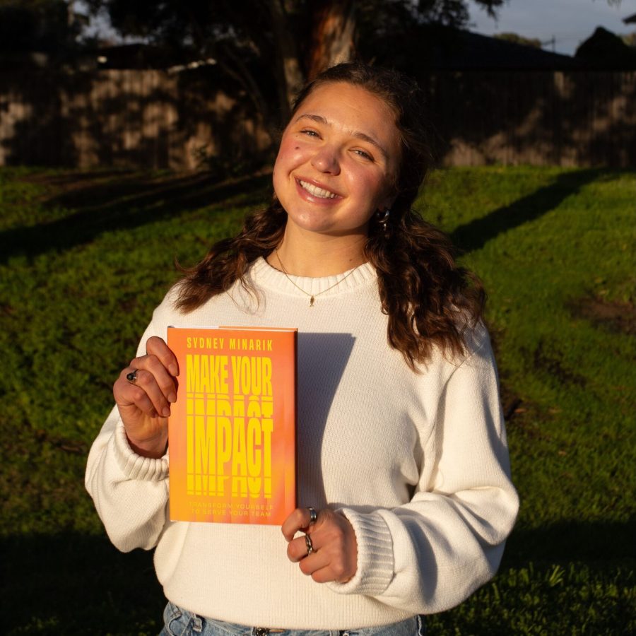 Sydney Minarik smiling outdoors holding her debut book Make Your Impact: Transform Yourself to Serve Your Team